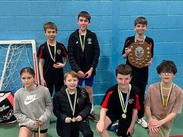 Seven children with medals, one holding a shield, posing inside a sports hall.