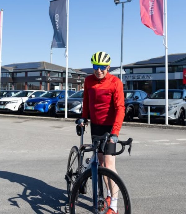 Cyclist in red jacket and yellow helmet standing with bike near parked cars.
