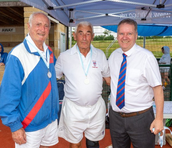 Three men smiling under a sports club tent, one wears a suit and two wear medals.