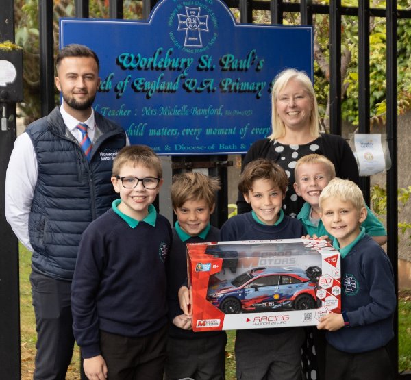 Group of children and two adults holding a toy car box outside a school gate.