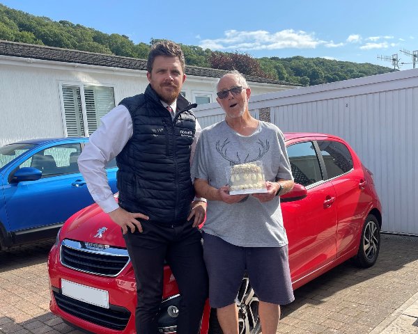 Two people standing by a red Peugeot; one is holding a cake.