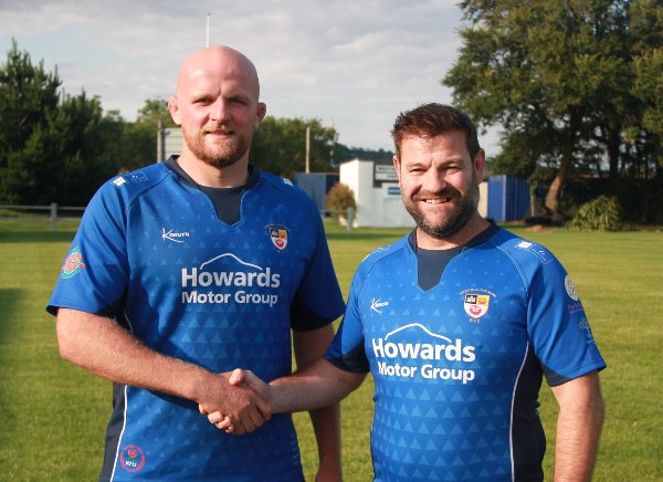 Two men in blue rugby kits shaking hands on a rugby pitch.