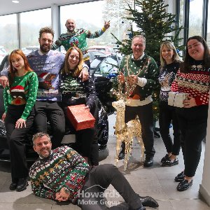 A group of people in Christmas jumpers gathered around a car with presents and a lit reindeer.