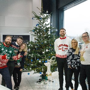 Group of people in Christmas jumpers standing by a decorated Christmas tree indoors.