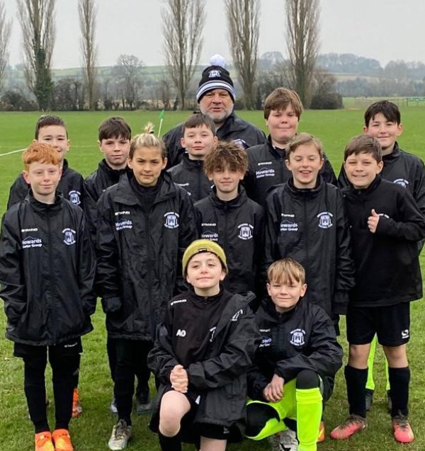 Youth football team with coach, wearing matching black jackets, posing on a grass field.