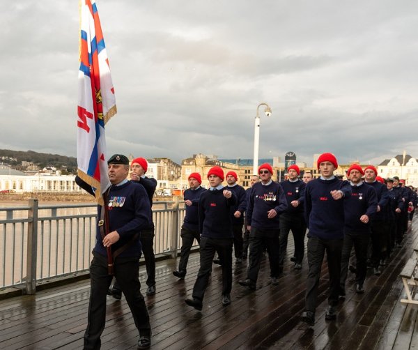 Group of people in navy uniforms marching on a boardwalk, one holding a flag.