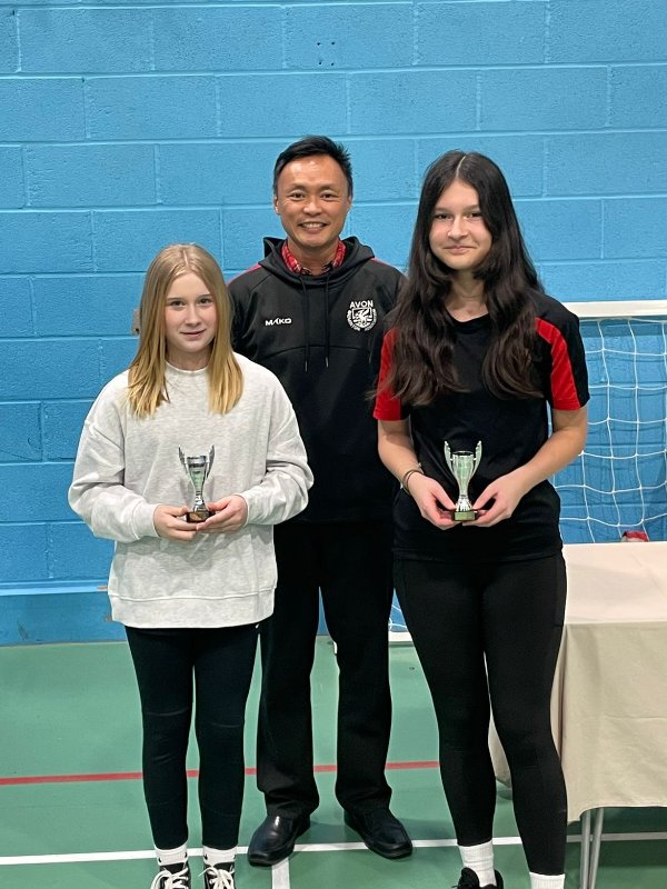 Two young people holding trophies with a smiling adult standing behind them.