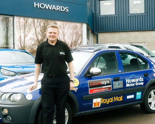 Man in black polo leaning on blue car with various logos, at Howards Motor Group building.