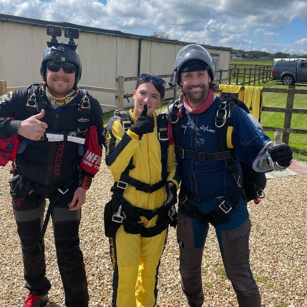 Three people in skydiving gear with parachutes, posing and smiling outdoors.