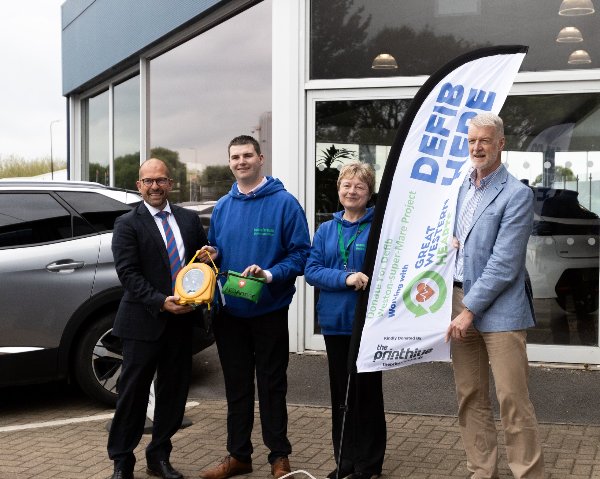 Four people standing outside Howards Motor Group with a defibrillator and a banner.