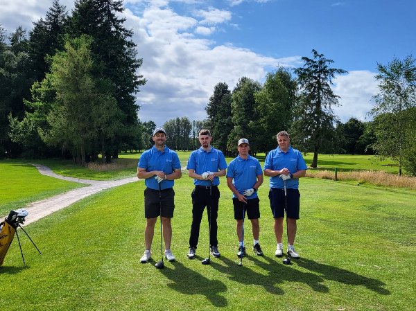 Four men in blue shirts standing on a golf course holding clubs.