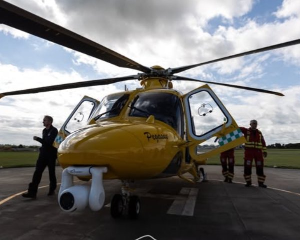 Yellow helicopter with open doors on a helipad, three people standing nearby.