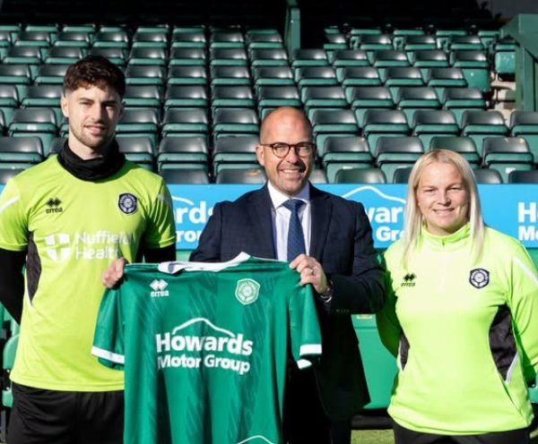 Three people holding a green football shirt with sponsor logos in a sports stadium.