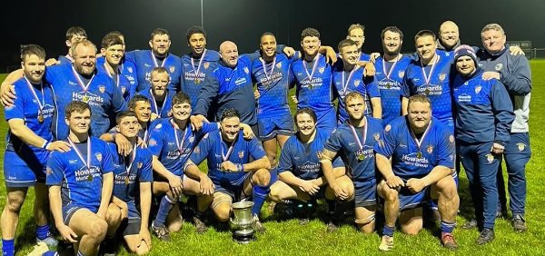 Rugby team in blue kits posing with a trophy on a field at night.