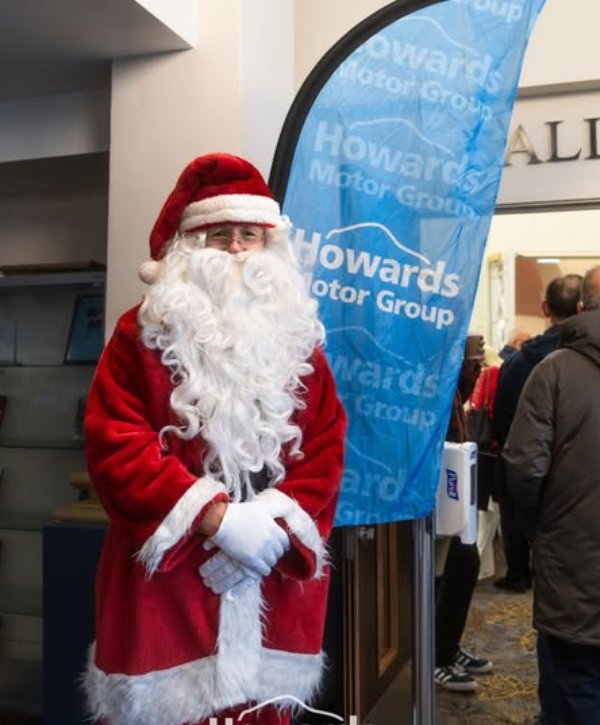 Santa Claus standing beside a Howards Motor Group banner indoors.