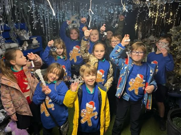 Group of children in blue Christmas jumpers holding decorations with lights above them.