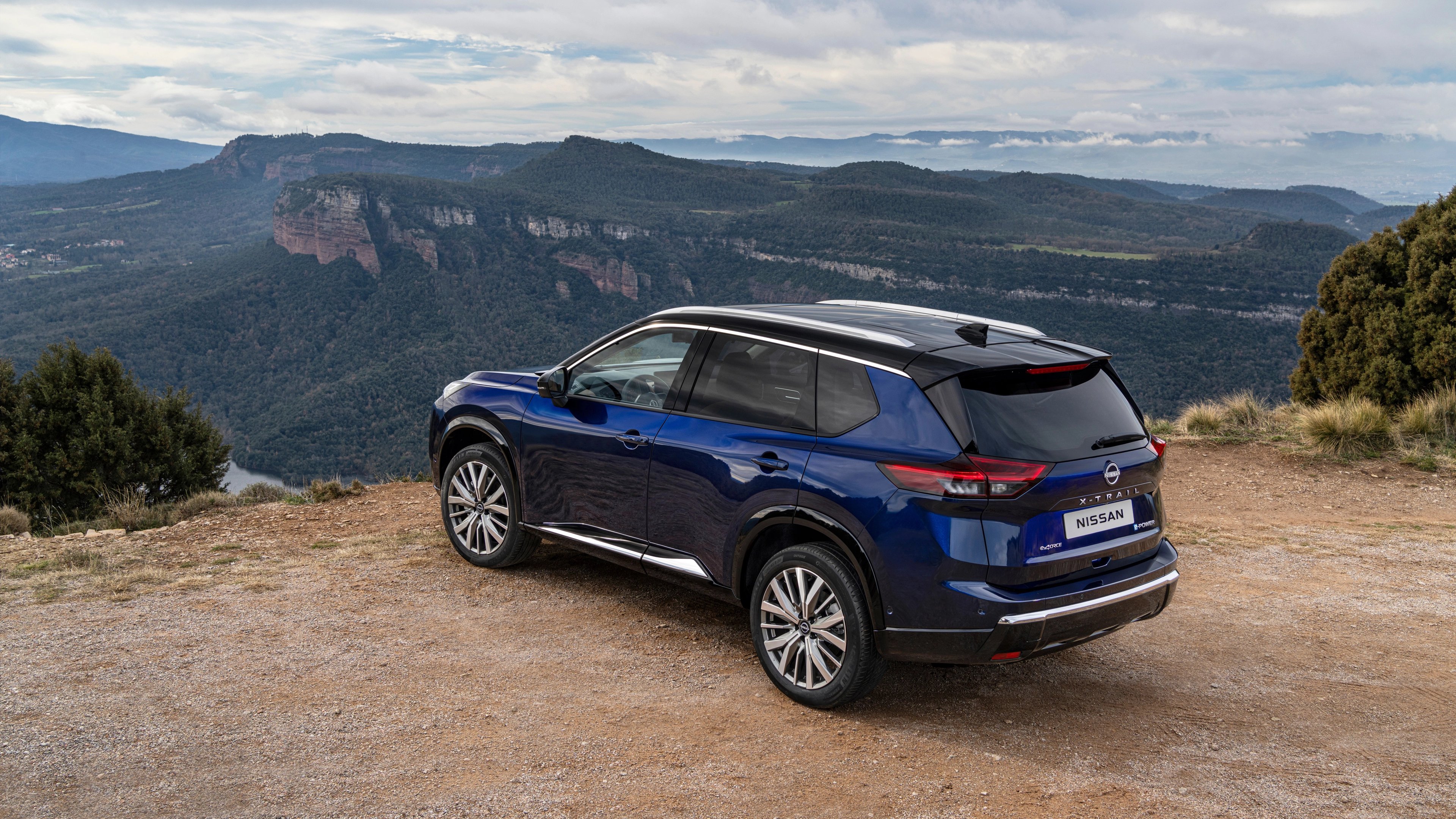 Blue Nissan X-Trail parked on a hilltop overlooking a scenic mountainous landscape.