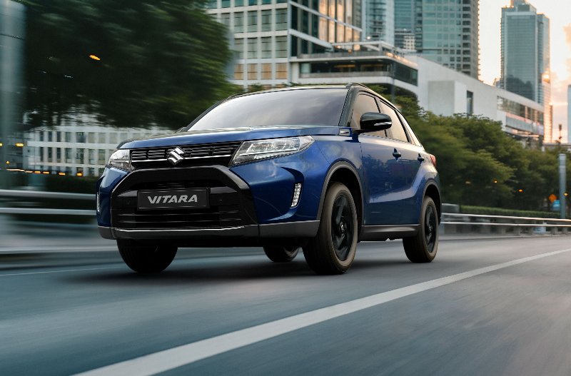 Blue Suzuki Vitara driving on an urban road with buildings in the background.