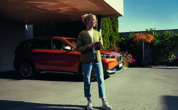 Person standing next to an orange car parked in a driveway.