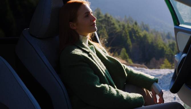 Person sitting in a car, wearing a green coat, with trees visible outside the window.