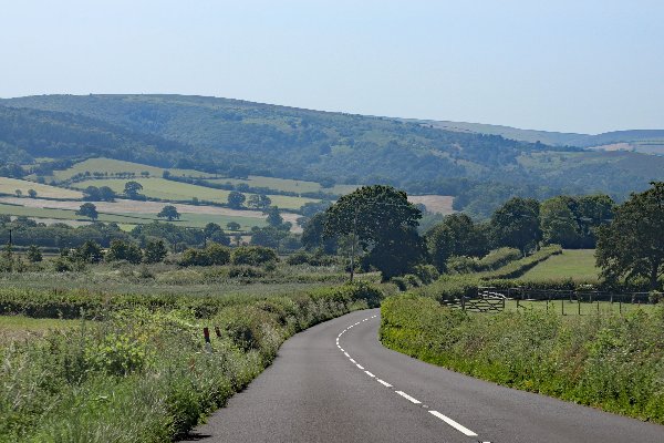 Curving country road surrounded by green fields and distant hills.