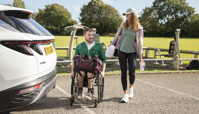 Man in wheelchair and woman walking by a car in a car park.