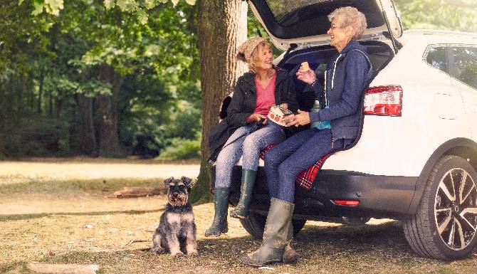 Two people sitting in an open car boot eating snacks, with a small dog nearby.