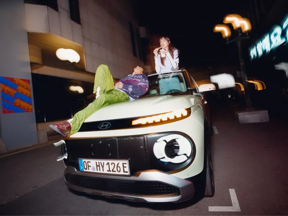 Two people on a parked car at night, one lying on the bonnet, the other sitting on the roof.