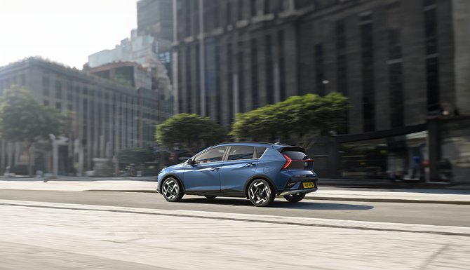 Blue hatchback car driving past city buildings.