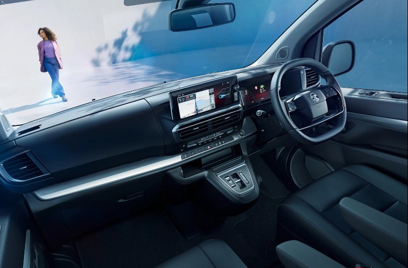 Car interior with dashboard displays, steering wheel, and a person walking past outside.