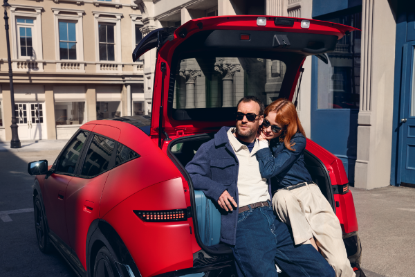 Couple sitting in the open boot of a red car parked by a building.