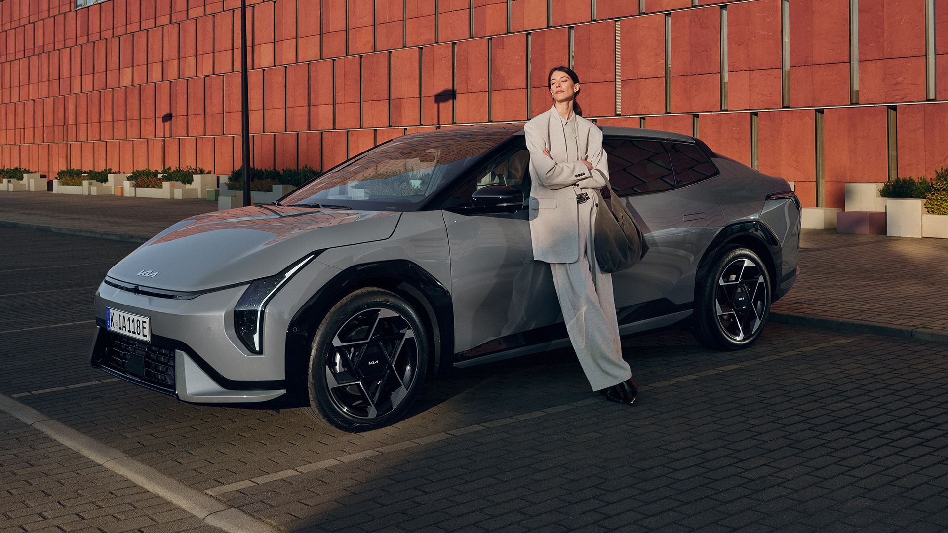 Person in grey coat leans against a grey car parked on a paved area.