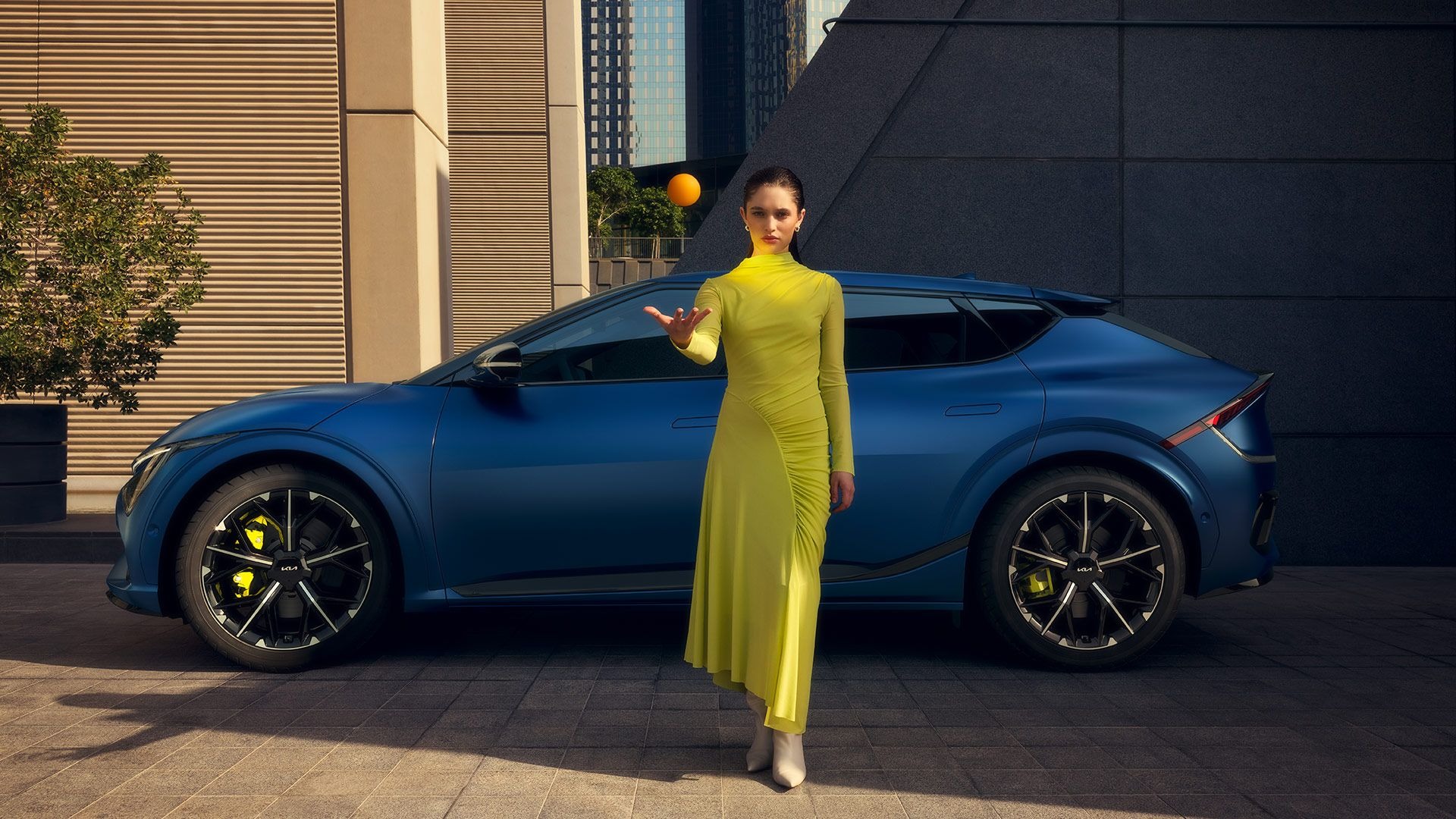 Woman in a yellow dress standing by a blue car, holding an orange ball above her hand.