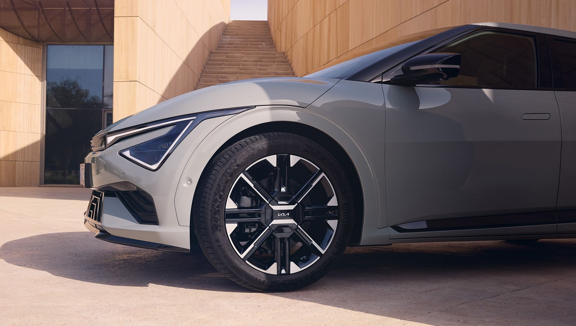 Close-up of a grey car's front wheel and headlight near modern building steps.