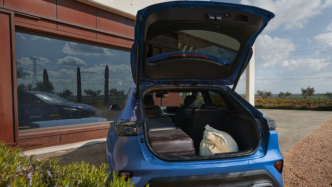Open boot of a blue car with luggage inside, parked beside a window reflecting trees and sky.