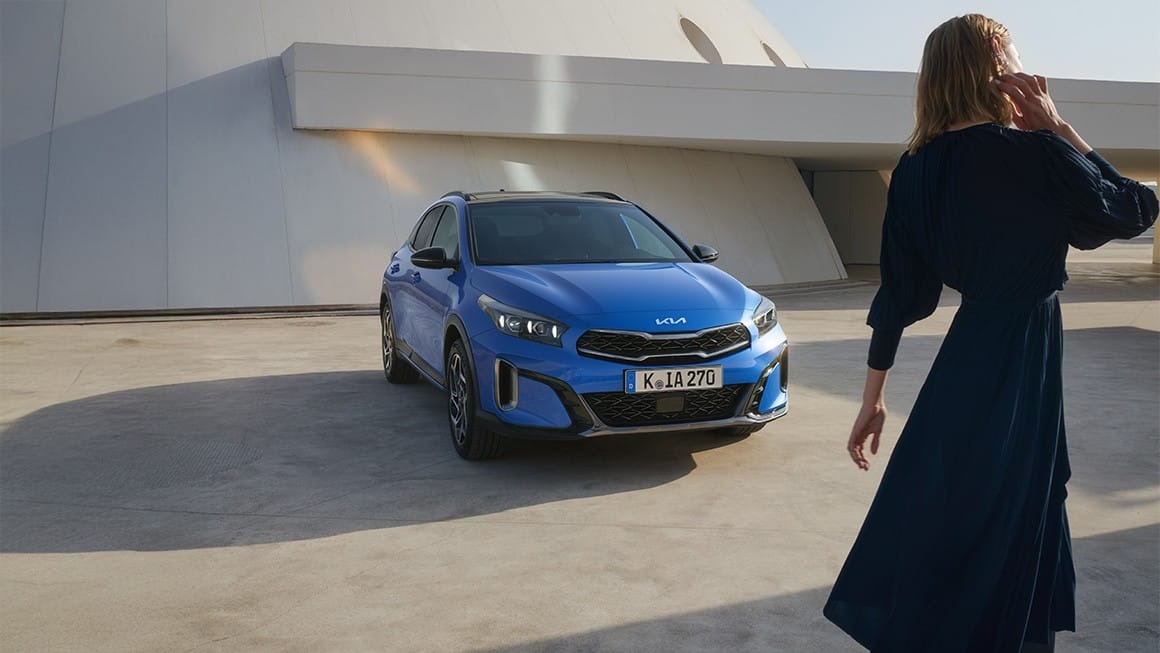 Blue Kia car parked with a person in a dark dress walking nearby.