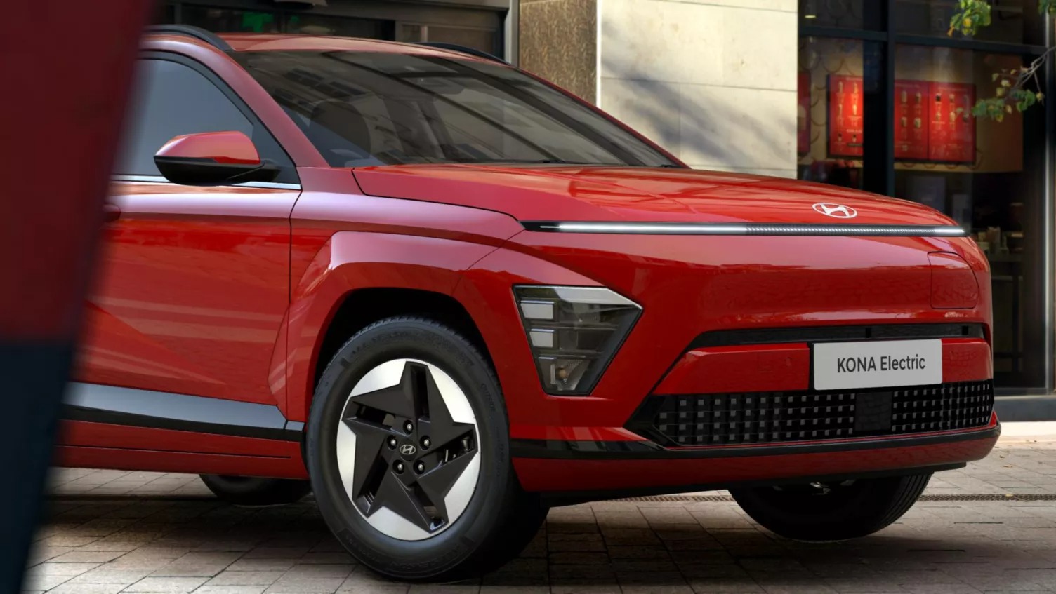 Red Hyundai Kona Electric car, front and side view, parked on a street.