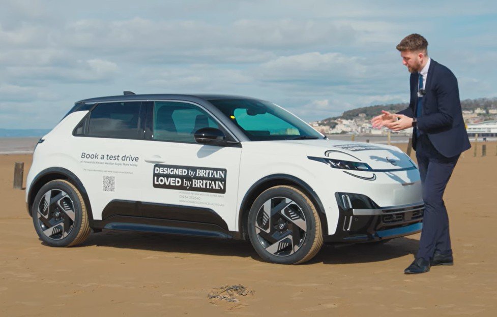 Man in suit standing next to a white car on a sandy beach.