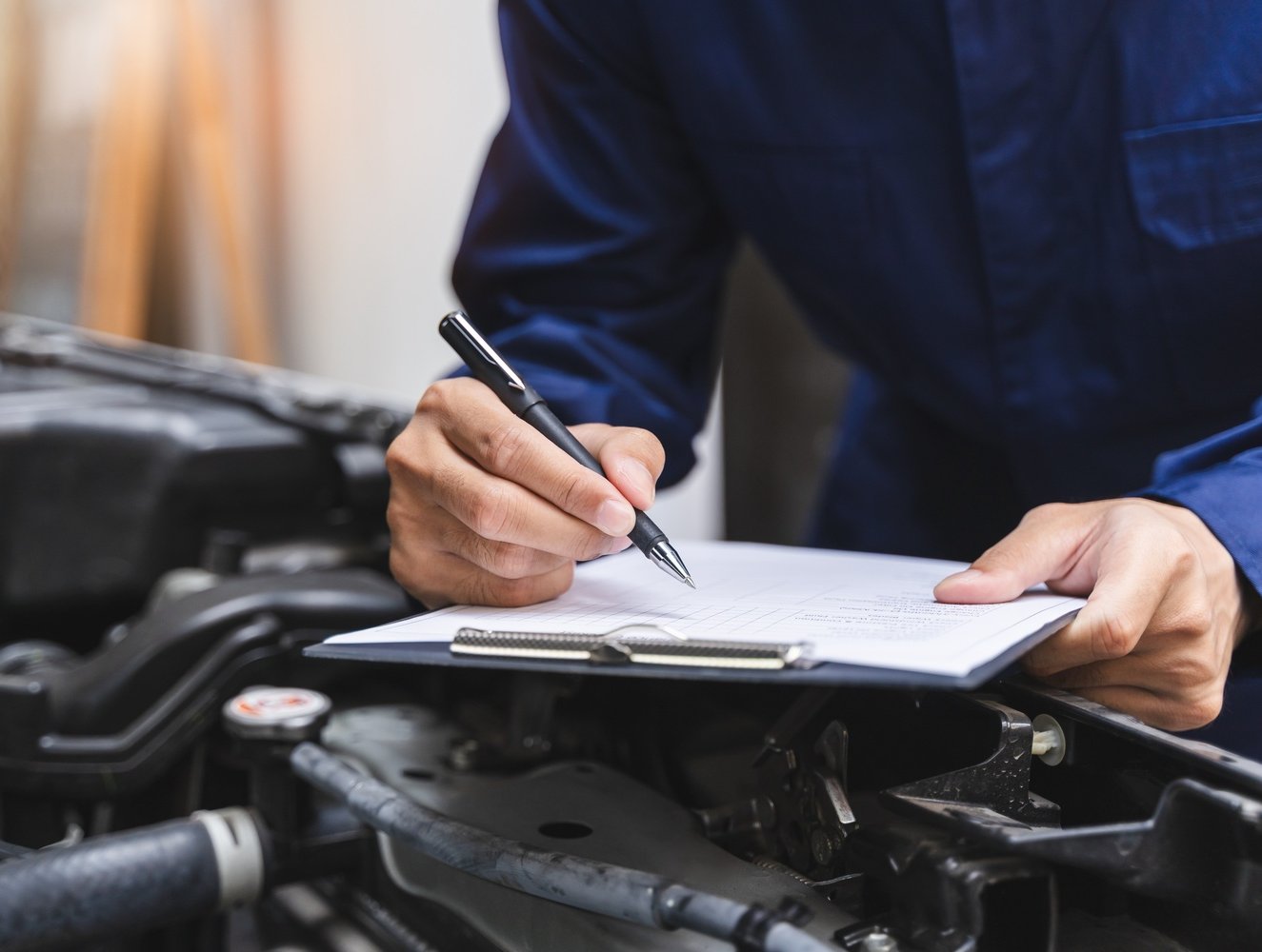 Mechanic writing on a clipboard next to a car engine.