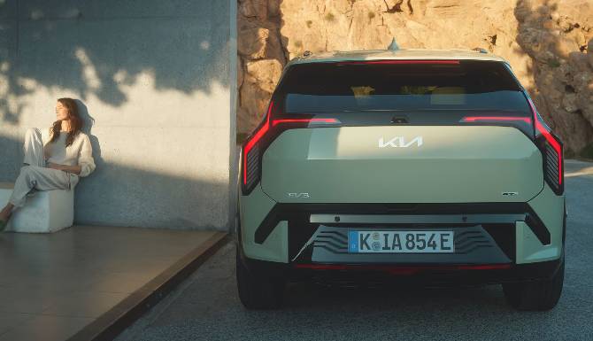 Rear view of a Kia EV9 parked next to a seated person on a concrete bench.