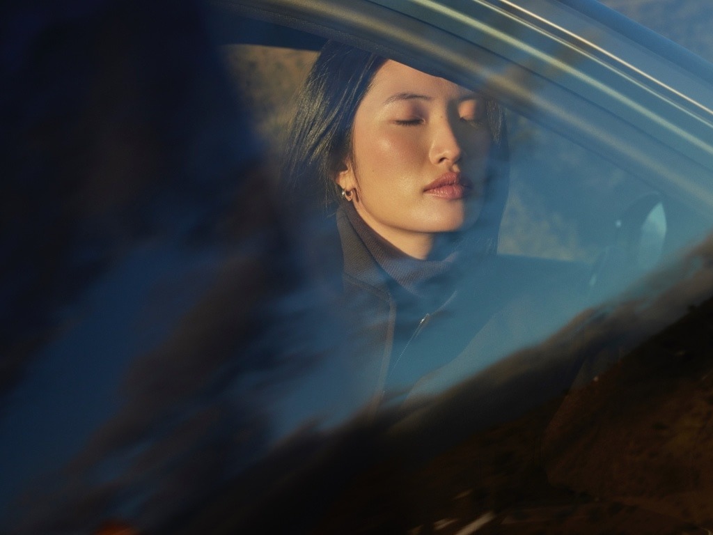 Person with closed eyes sitting in a car, viewed through a window.