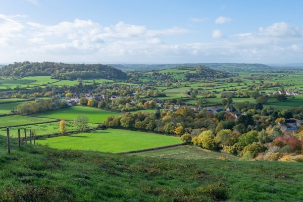 View of a countryside village surrounded by green fields and trees under a partly cloudy sky.