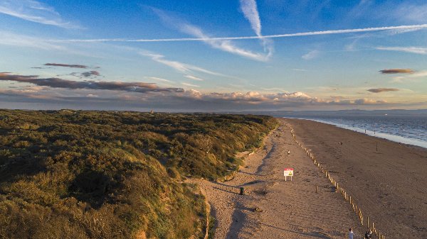 Sandy beach with people walking, bordered by grassy dunes, under a sky with streaked clouds.