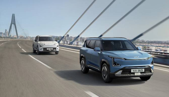 Two modern SUVs driving on a bridge road; blue SUV in front, white SUV behind.