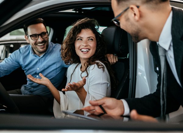 Three people smiling and talking inside a car, with a man holding a tablet outside the window.