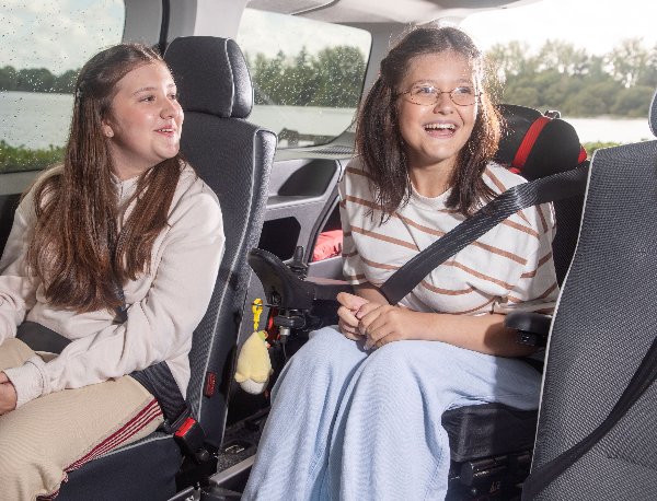 Two girls sitting inside a car, smiling and wearing seatbelts.
