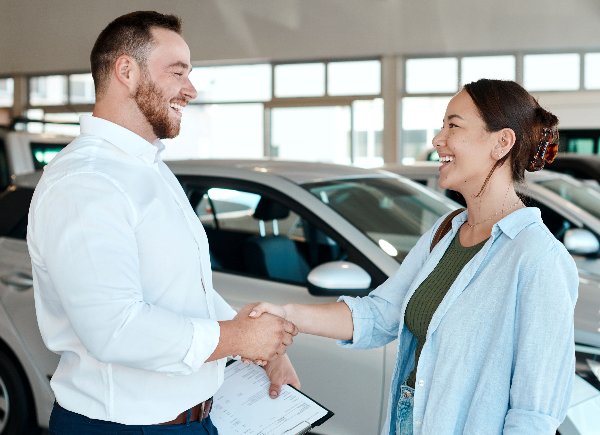 Two people shaking hands in a car showroom, one holding paperwork.