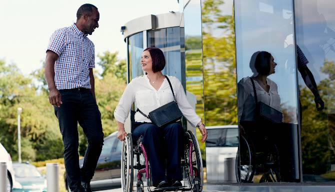 A person walking and a person in a wheelchair conversing outside a modern building.