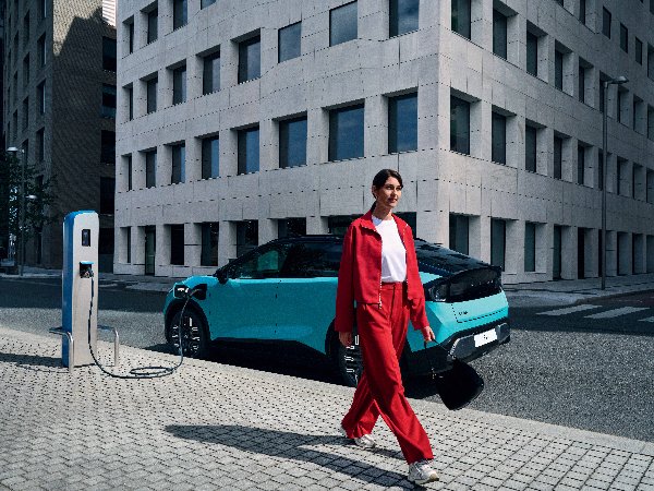 Woman in red outfit walking past a blue electric car charging in a city street.